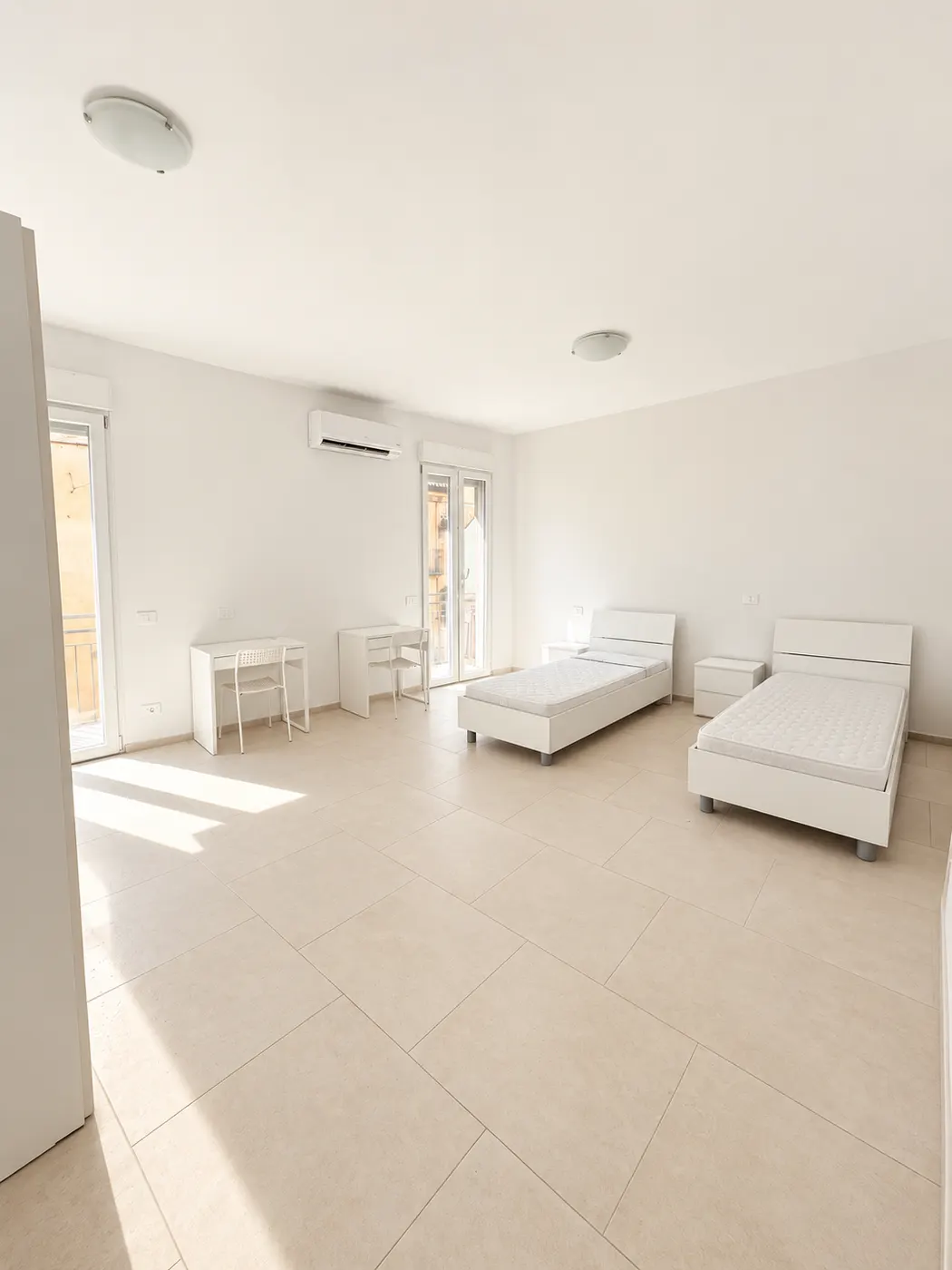 Spazio abitativo – Ambiente con due letti, scrivanie e accesso al balcone.
